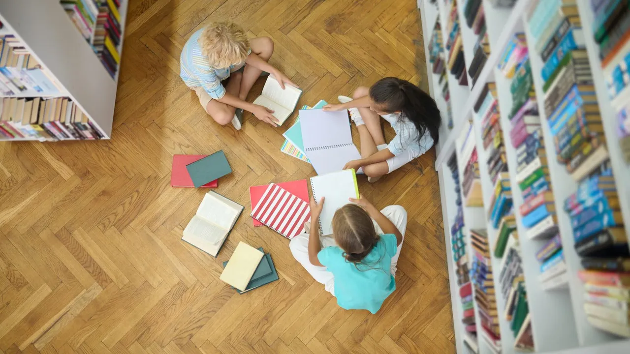 Três crianças sentadas no piso de madeira de uma biblioteca, vistas de cima, com livros e cadernos abertos entre as estantes