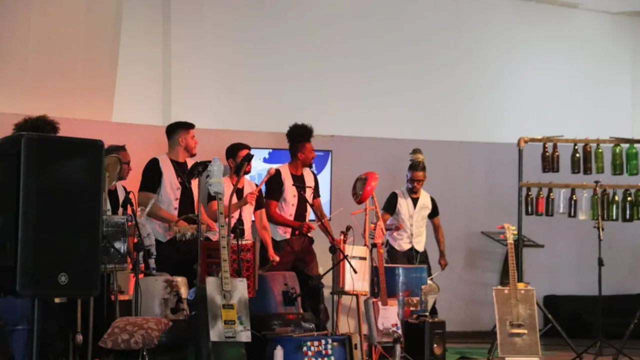 Músicos da Orquestra de Sucata no palco com instrumentos construídos com materiais reaproveitados, como latas, garrafas e tambores